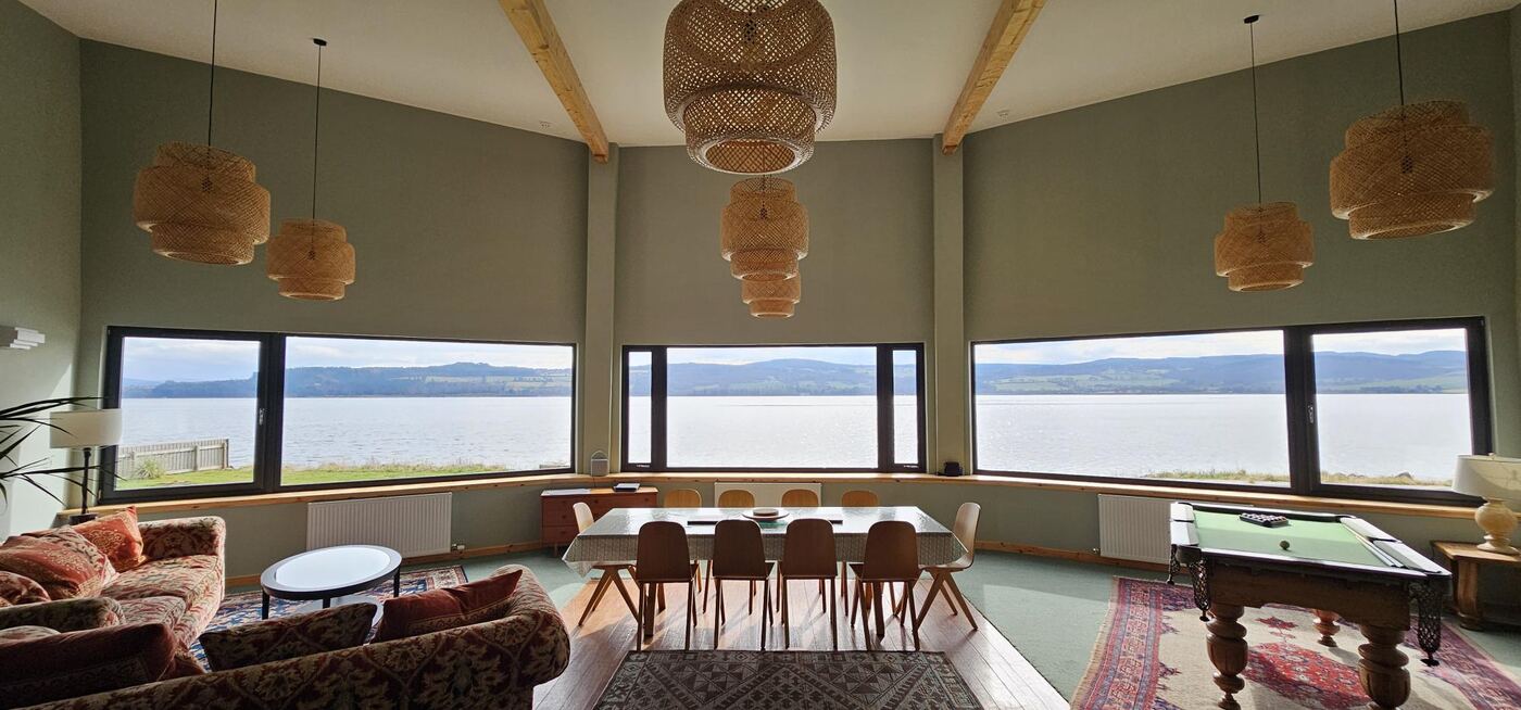 Finished interior living space with wood-beam ceiling, woven pendant lights and a full run of windows over the loch