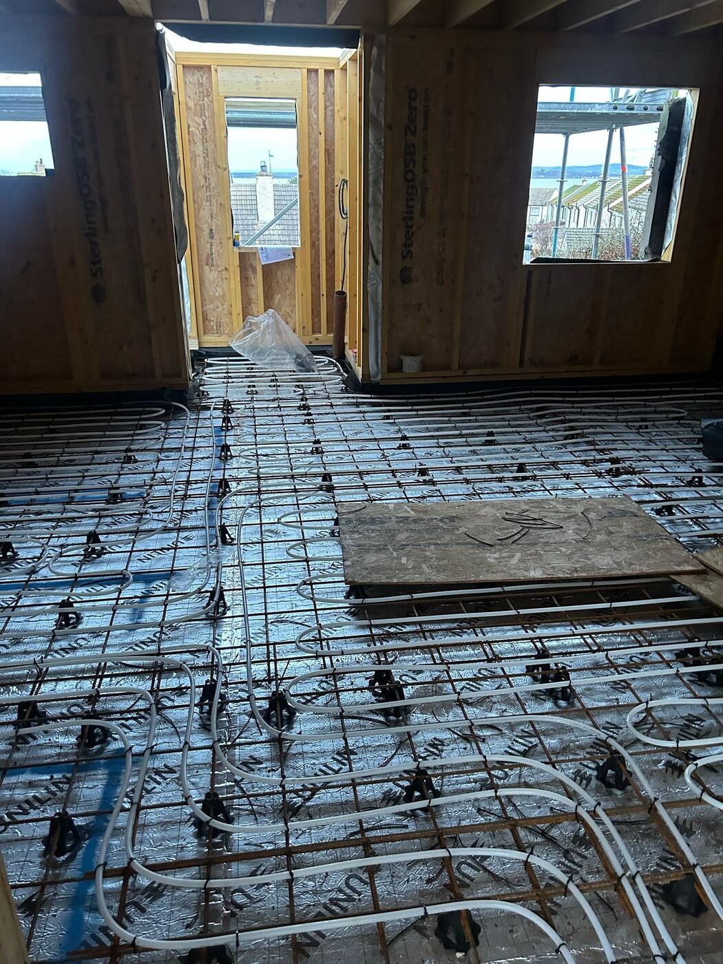 Underfloor heating pipework running through an open doorway between two rooms, with a view out to the water beyond